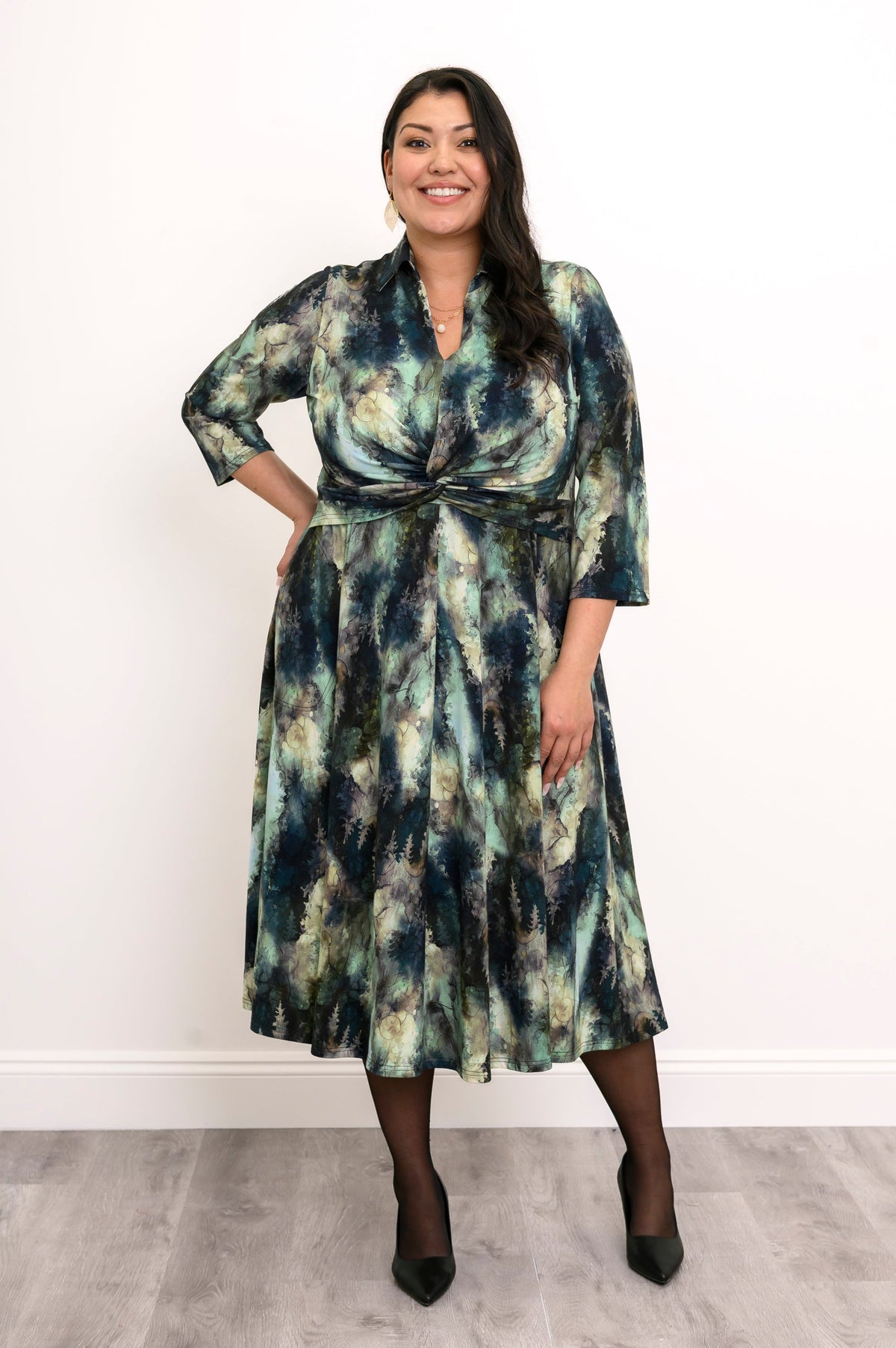 Front view of plus sized female model in a green forest print midi dress with a twisted waist, sheer black tights, and black heels, hand on hip, against a white wall and gray floor