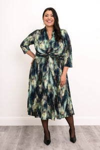 Front view of plus sized female model in a green forest print midi dress with a twisted waist, sheer black tights, and black heels, hand on hip, against a white wall and gray floor