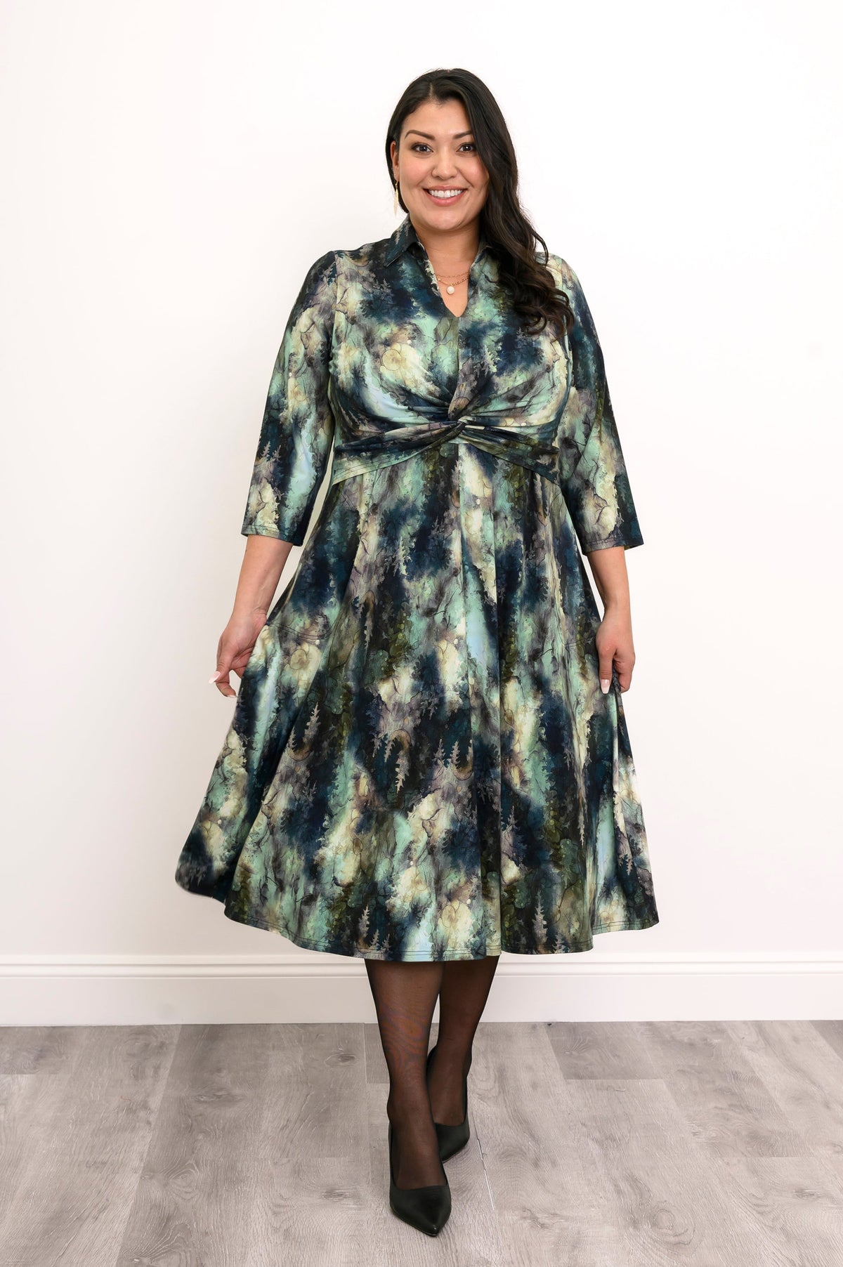 Front view of plus sized female model wearing a A-line watercolor tree pattern bamboo dress featuring a front twist, collar, and 3/4 sleeves, styled with heels and tights, against a white wall and gray floor