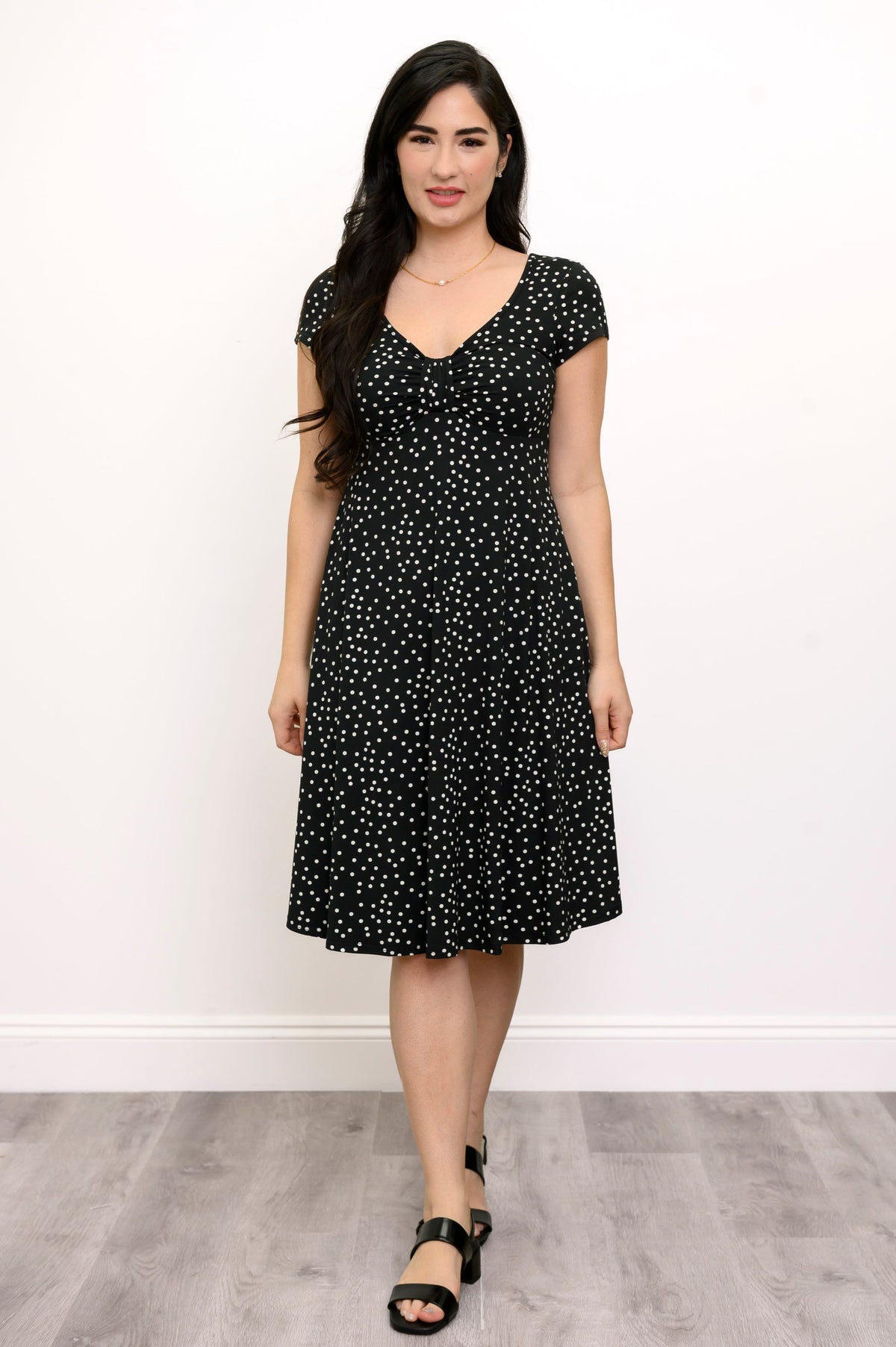 Woman wearing a short sleeve black dress with white polka dots and lack sandals against a white background