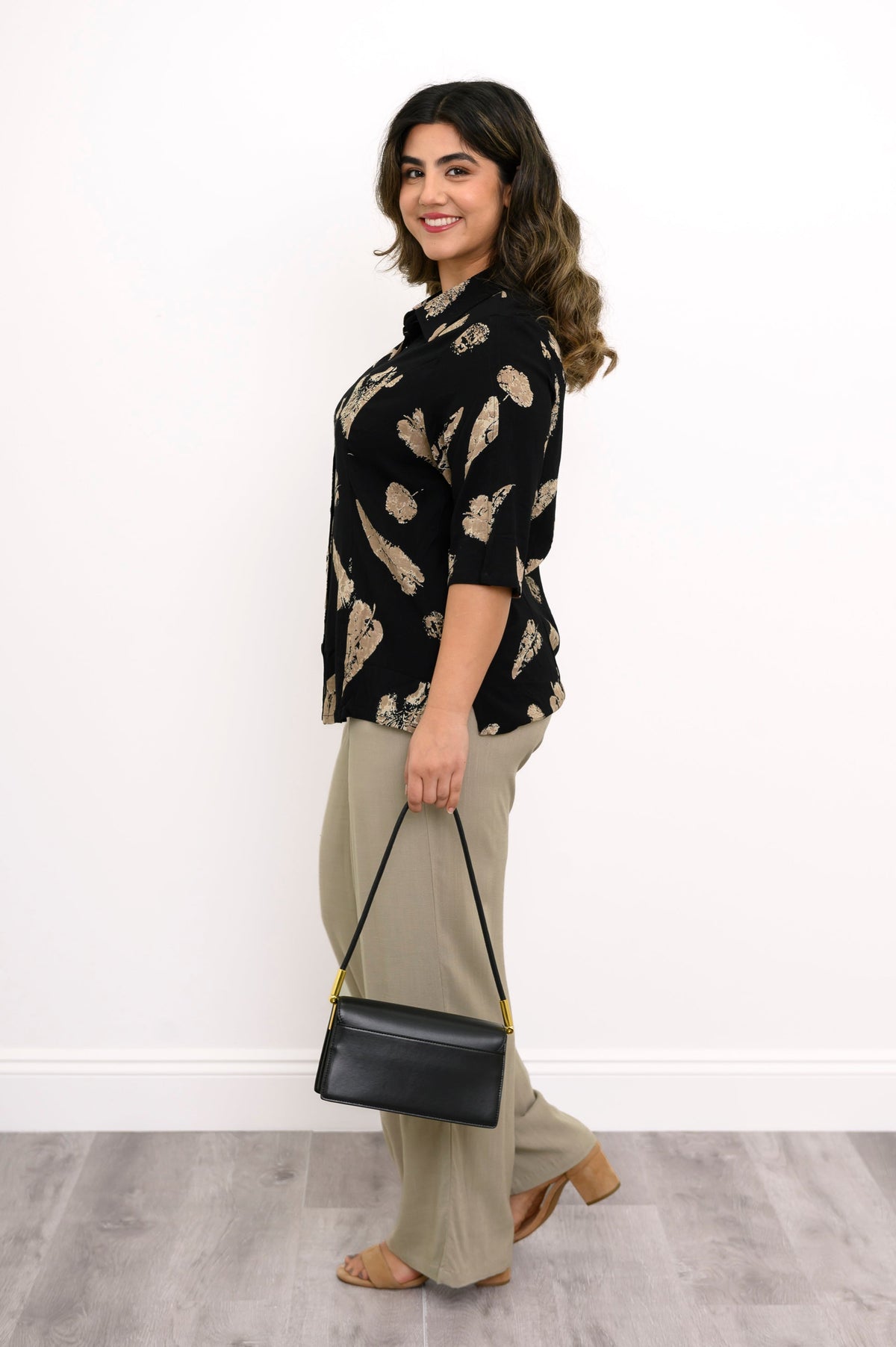 side view of woman wearing a black blouse with tan leaf pattern and beige pants styled with a black bag