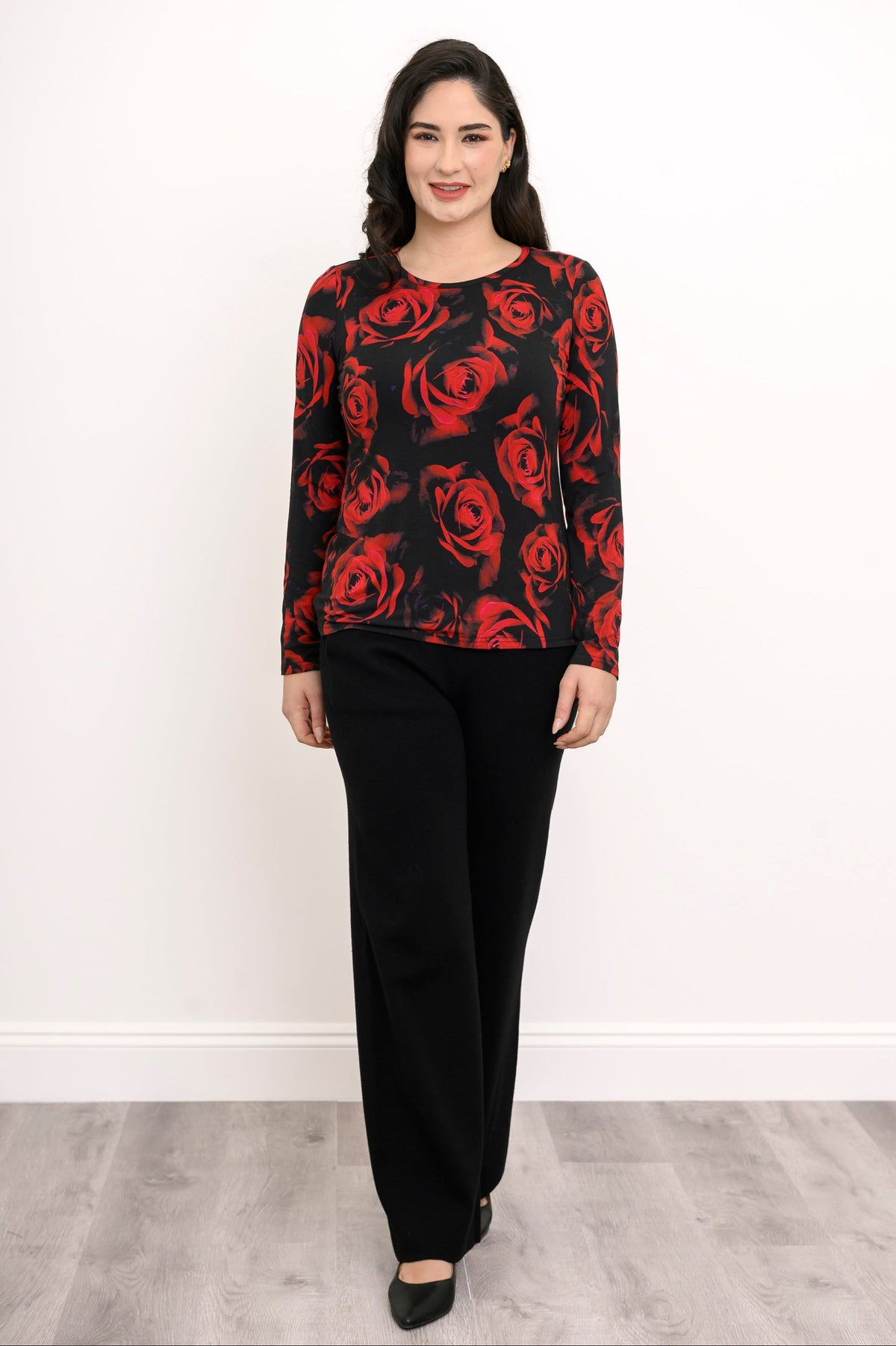 Front view of a female model wearing a long sleeve black top with red roses with black pants and heels, against a white wall and gray floor
