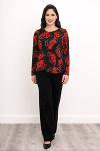 Front view of a female model wearing a long sleeve black top with red roses with black pants and heels, against a white wall and gray floor