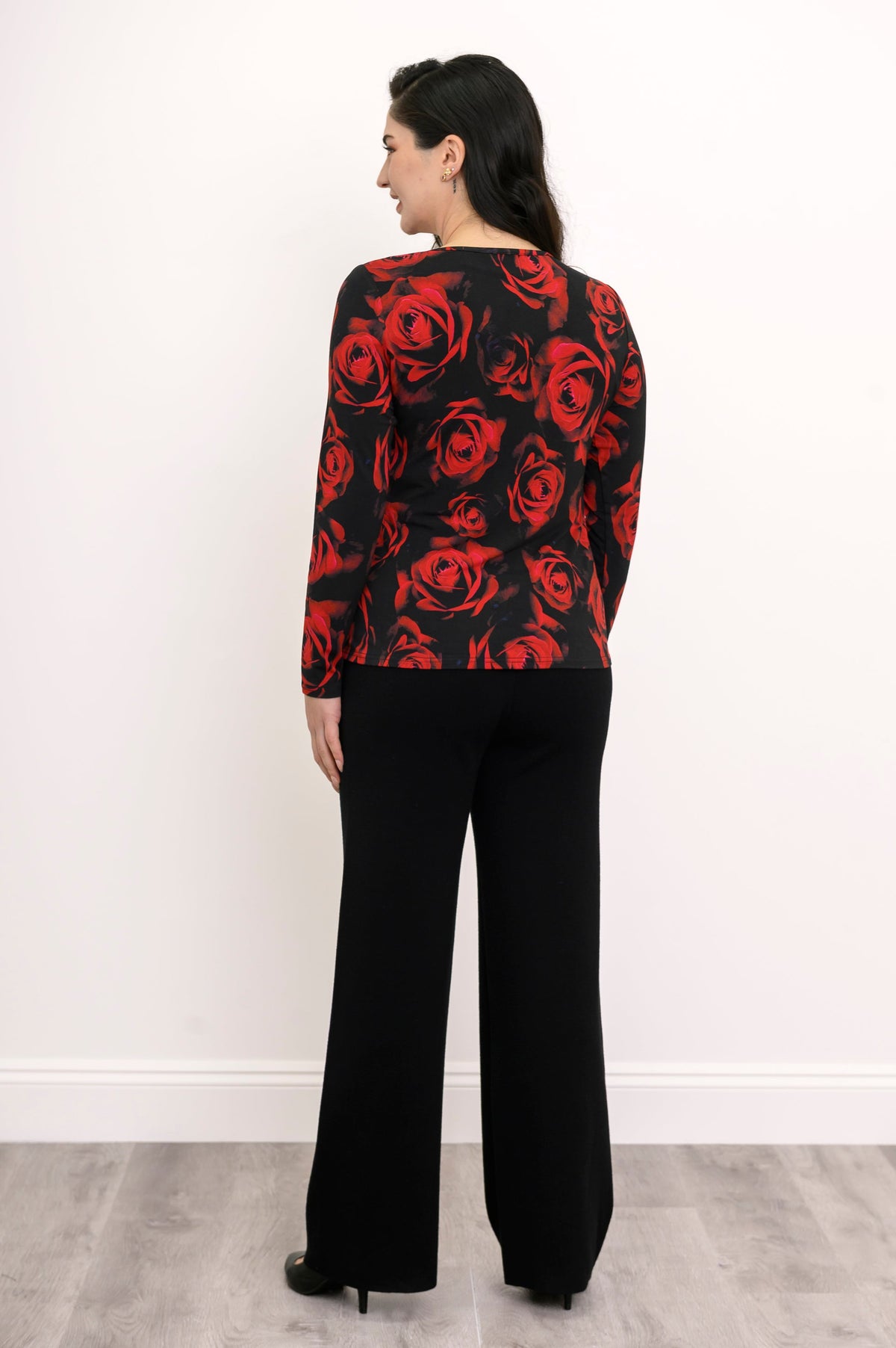 Back view of a female model wearing a long sleeve black top with red roses with black pants and heels, against a white wall and gray floor