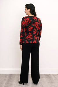 Back view of a female model wearing a long sleeve black top with red roses with black pants and heels, against a white wall and gray floor