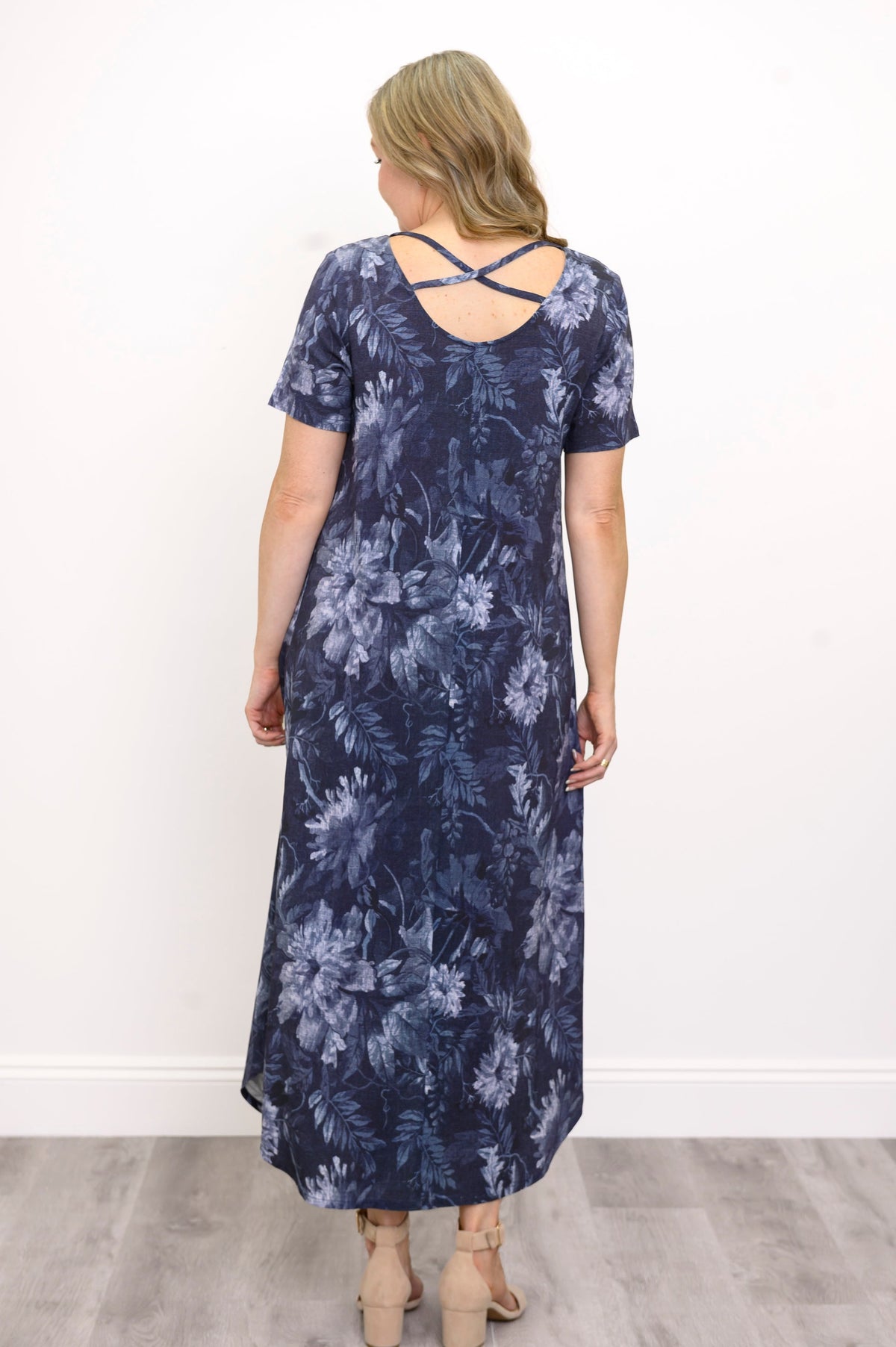 Back view of woman wearing long navy blue floral dress with pockets and sandals on a white background