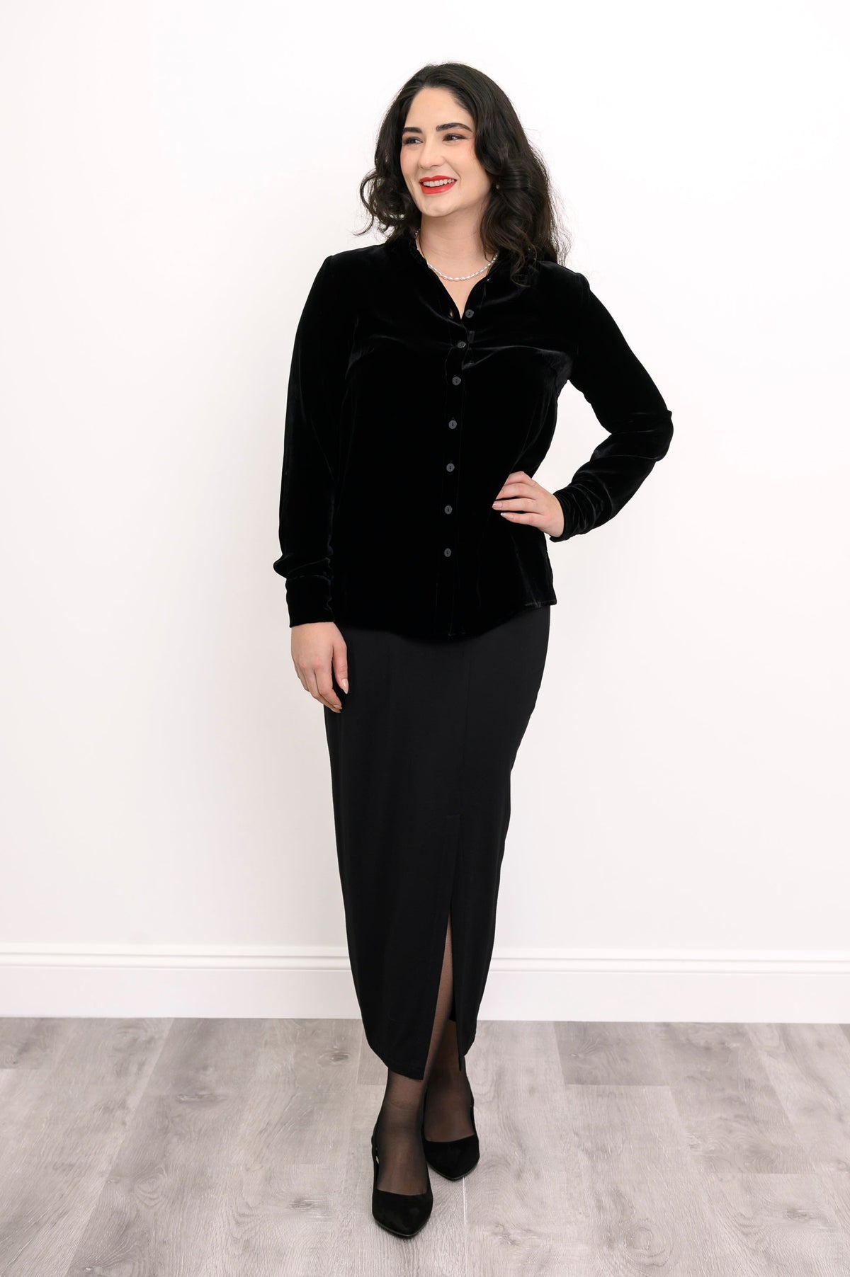 Wide studio shot of an elegant women's black velvet long-sleeve blouse paired with a long black skirt and heels
