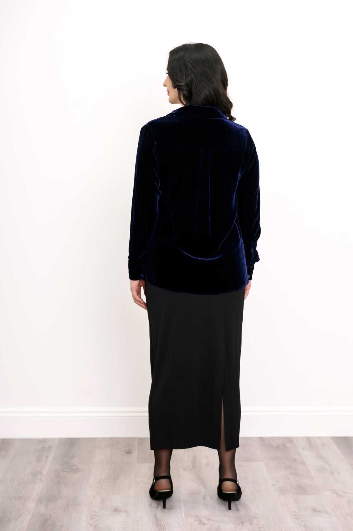 Back view of a woman wearing a navy blue silk velvet button-up shirt with a pleated back yoke and a black skirt