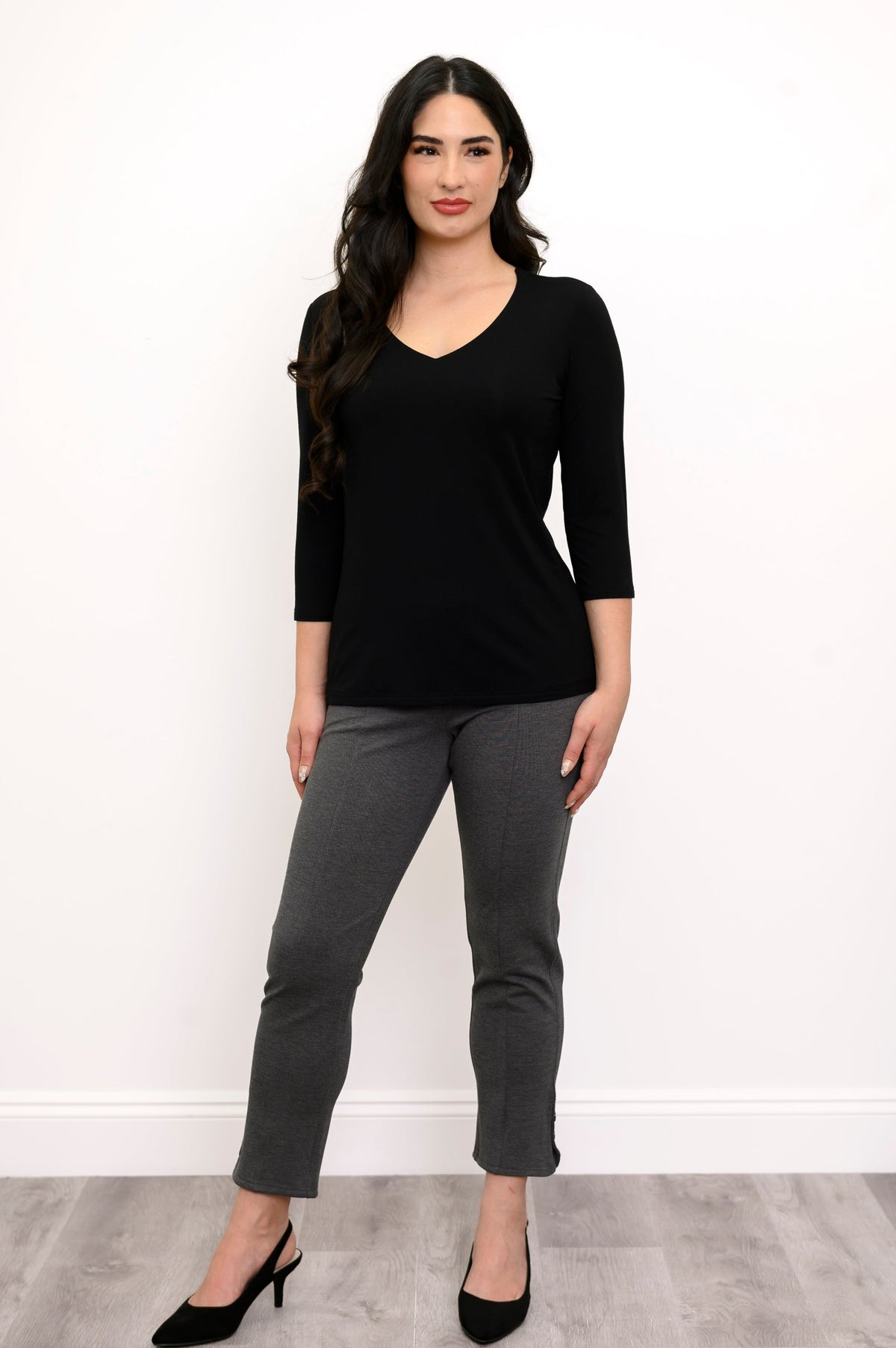 Full body view of woman with her hands at her side wearing a black top and grey slim fit pants