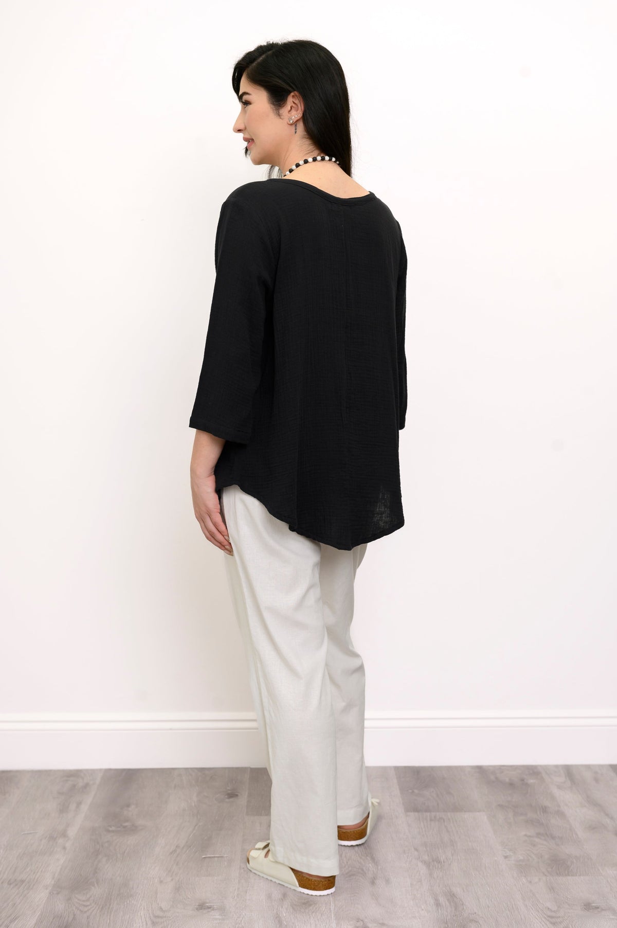 Woman wearing a black top and white pants with a beaded necklace against a white wall, back view