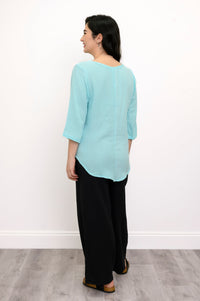 Woman wearing a light blue top, black pants, and black sandals against a white background, back view