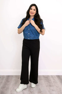 Model smiling in a cropped blue denim jacket with hands on collar and black pants