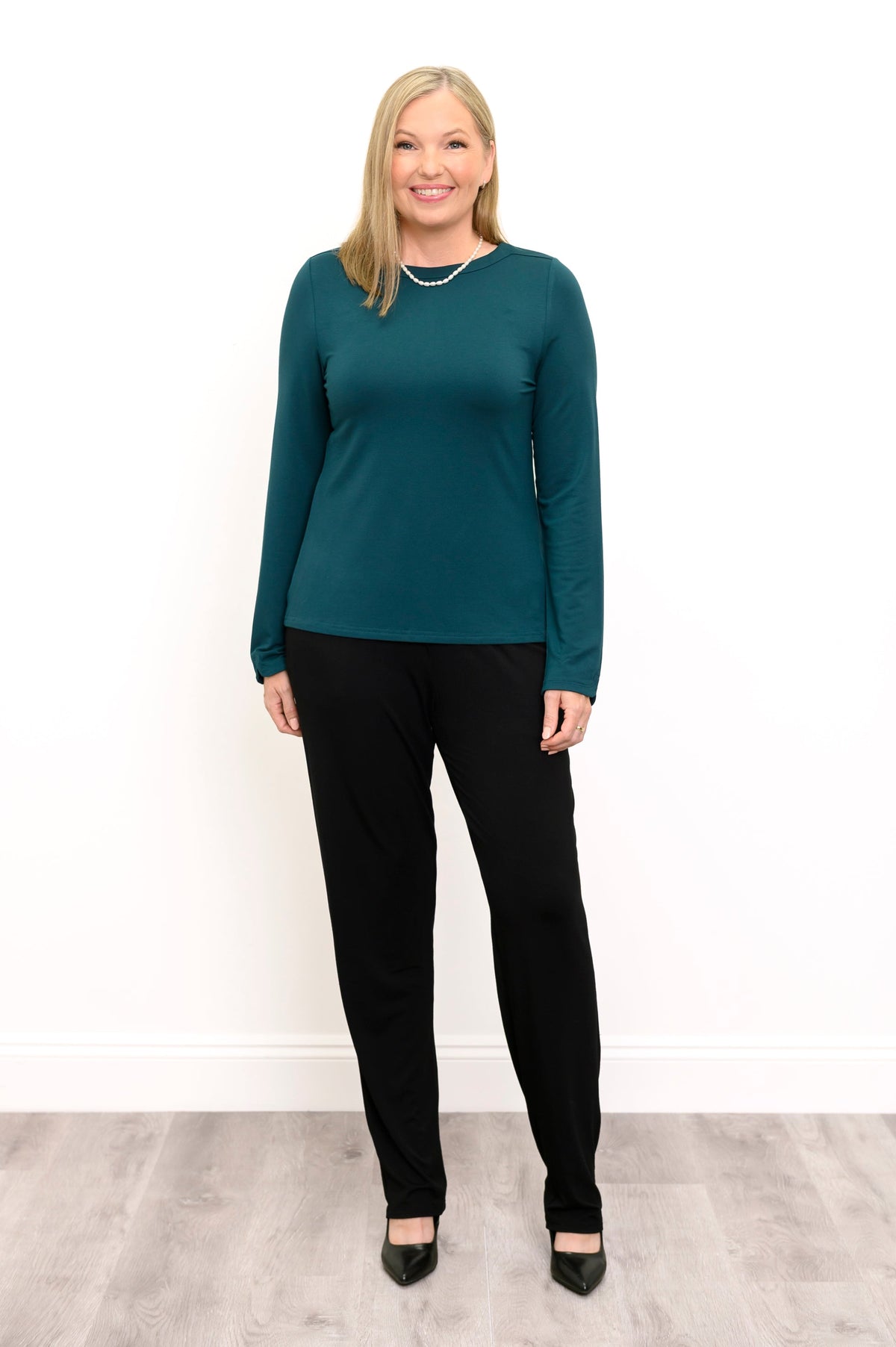 Front view of a female model wearing a teal long sleeve top with black pants and shoes, against a white wall and gray floor