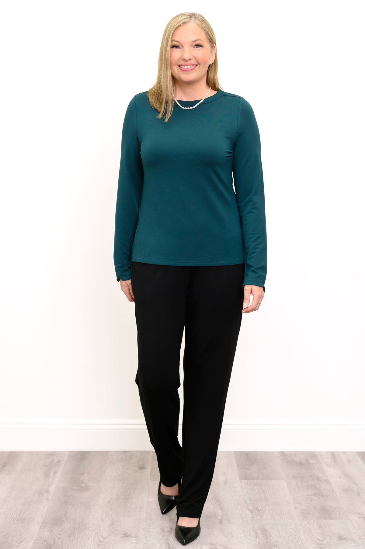 Front view of a female model in walking pose wearing a teal long sleeve top with black pants and shoes, against a white wall and gray floor
