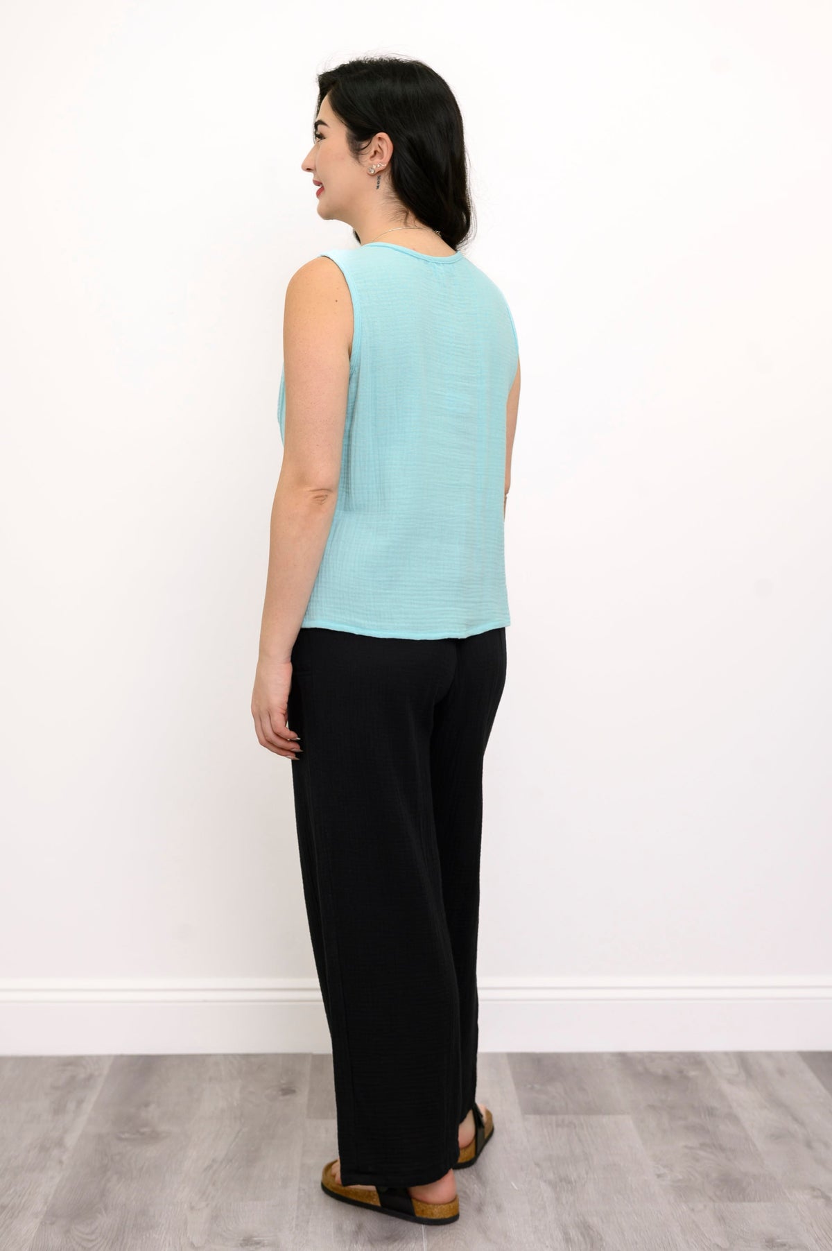 Woman wearing a light blue sleeveless top, black pants and black sandals  against a white wall, back view