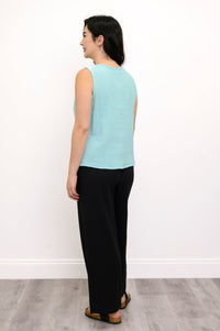 Woman wearing a light blue sleeveless top, black pants and black sandals  against a white wall, back view