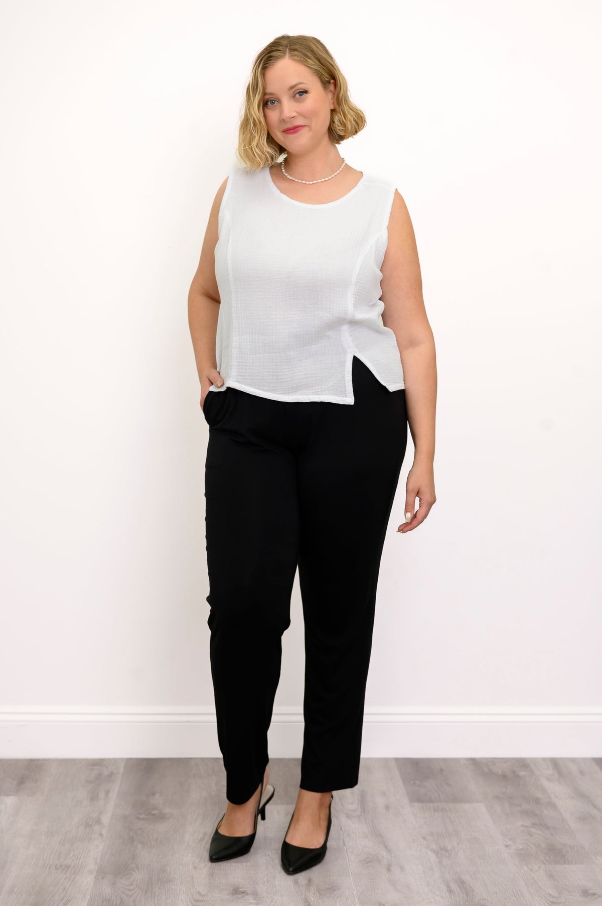 Woman wearing a white sleeveless top, black pants, and black shoes on a white background, front view