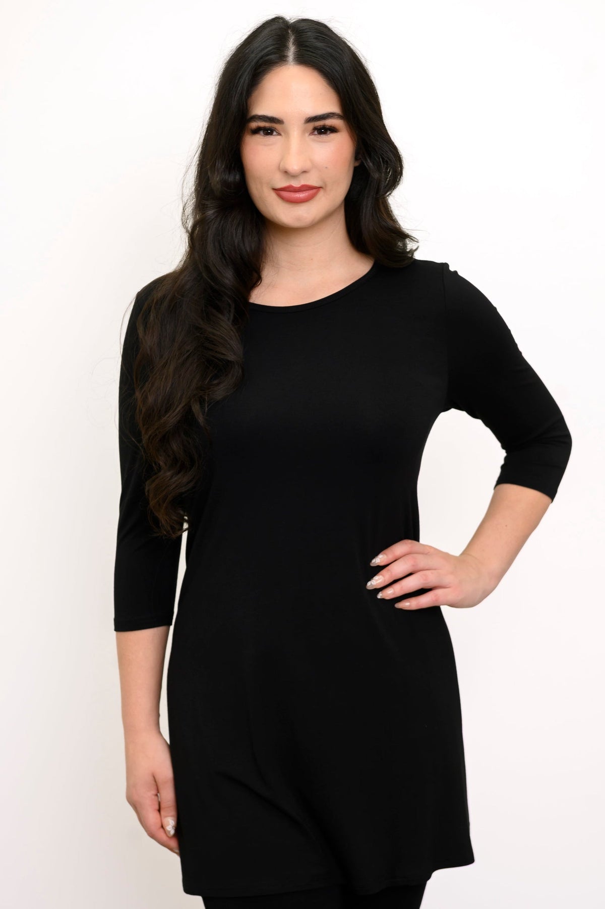 Close up front view of woman wearing long sleeve black tunic, smiling with her hand on her hip