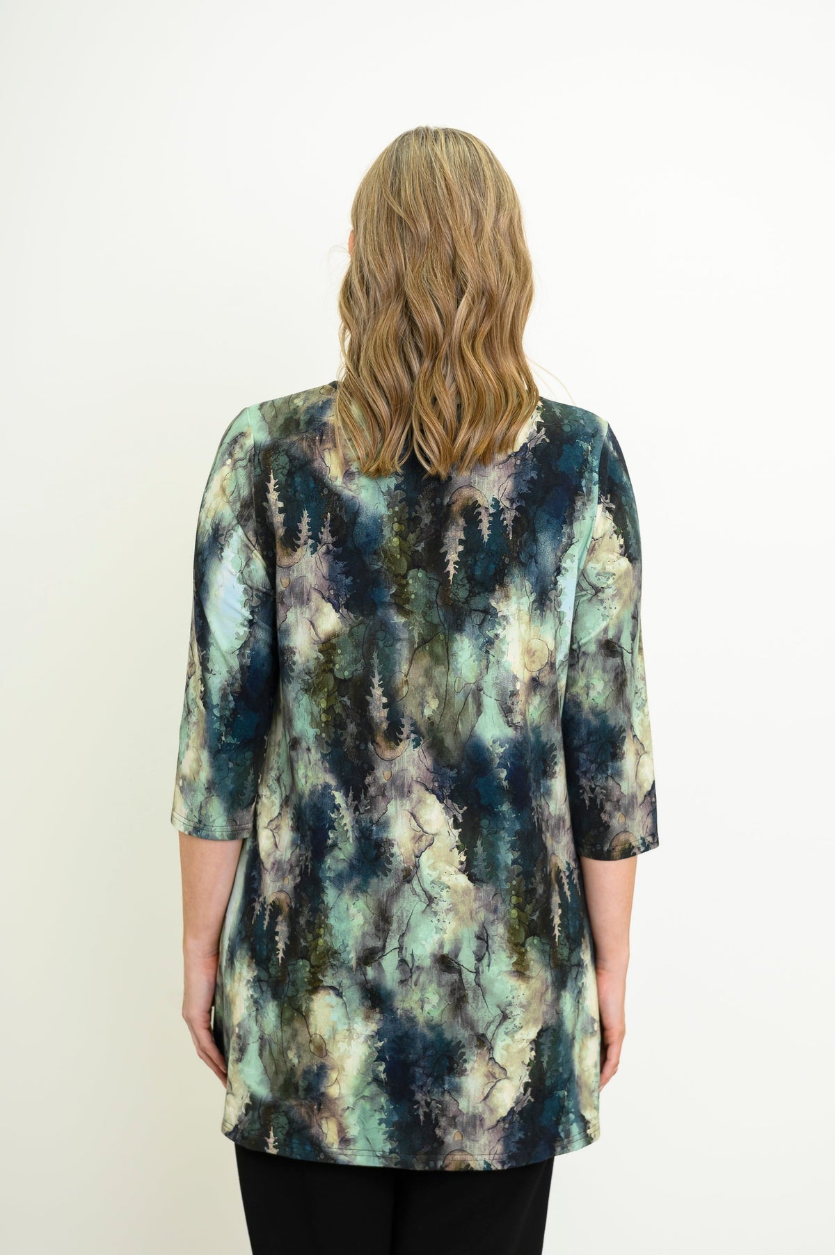 Rear view of a a female model wearing a blue-green watercolor forest print A-line tunic with 3/4 sleeves with black pants. Against a white backdrop