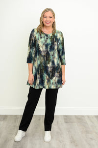 Front view of a female model wearing a blue-green watercolor forest print A-line tunic with 3/4 sleeves with black pants and white shoes. Against a white wall and gray floor