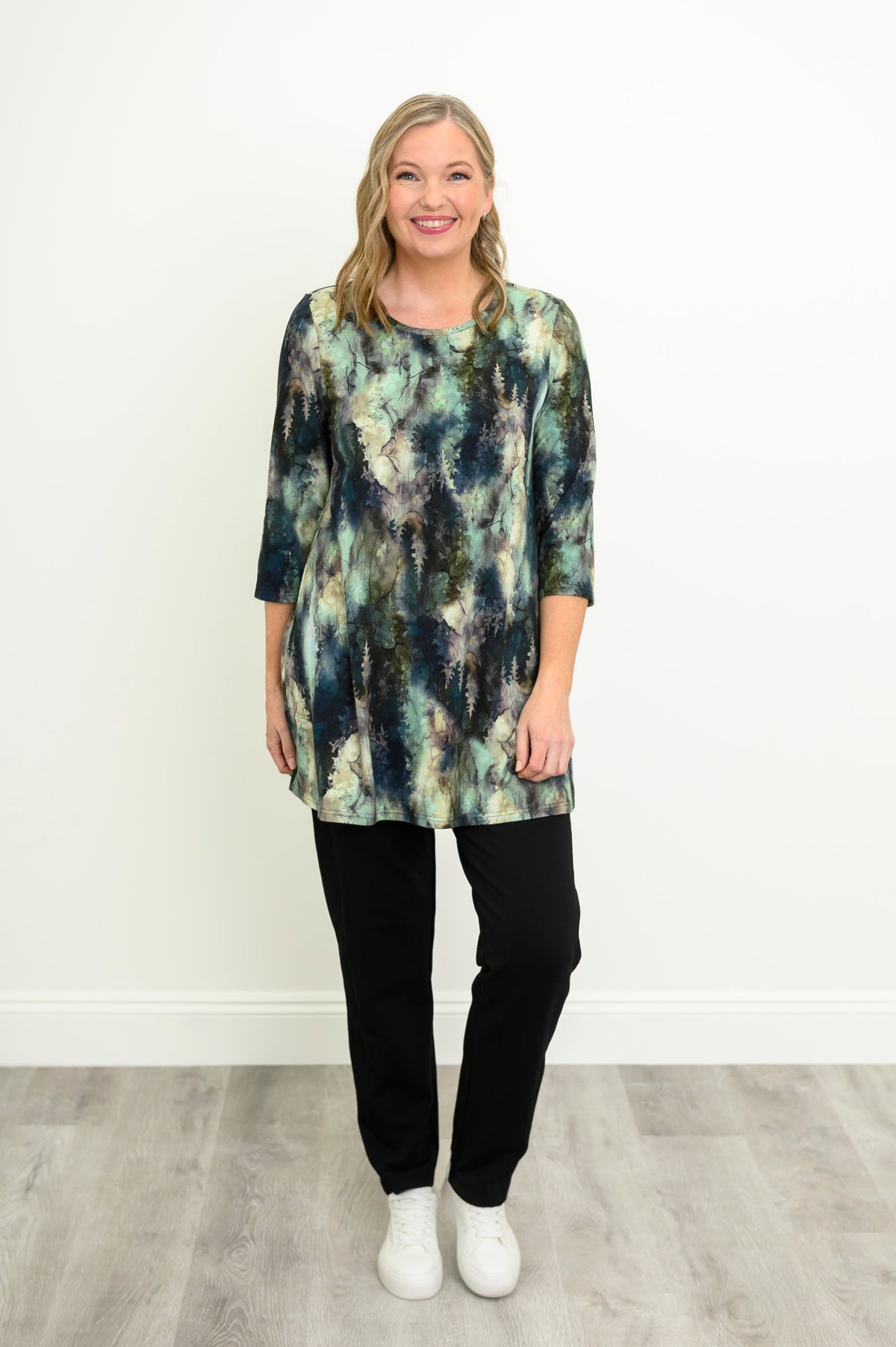 Front view of a female model wearing a blue-green watercolor forest print A-line tunic with 3/4 sleeves with black pants and white shoes, one leg propped up. Against a white wall and gray floor
