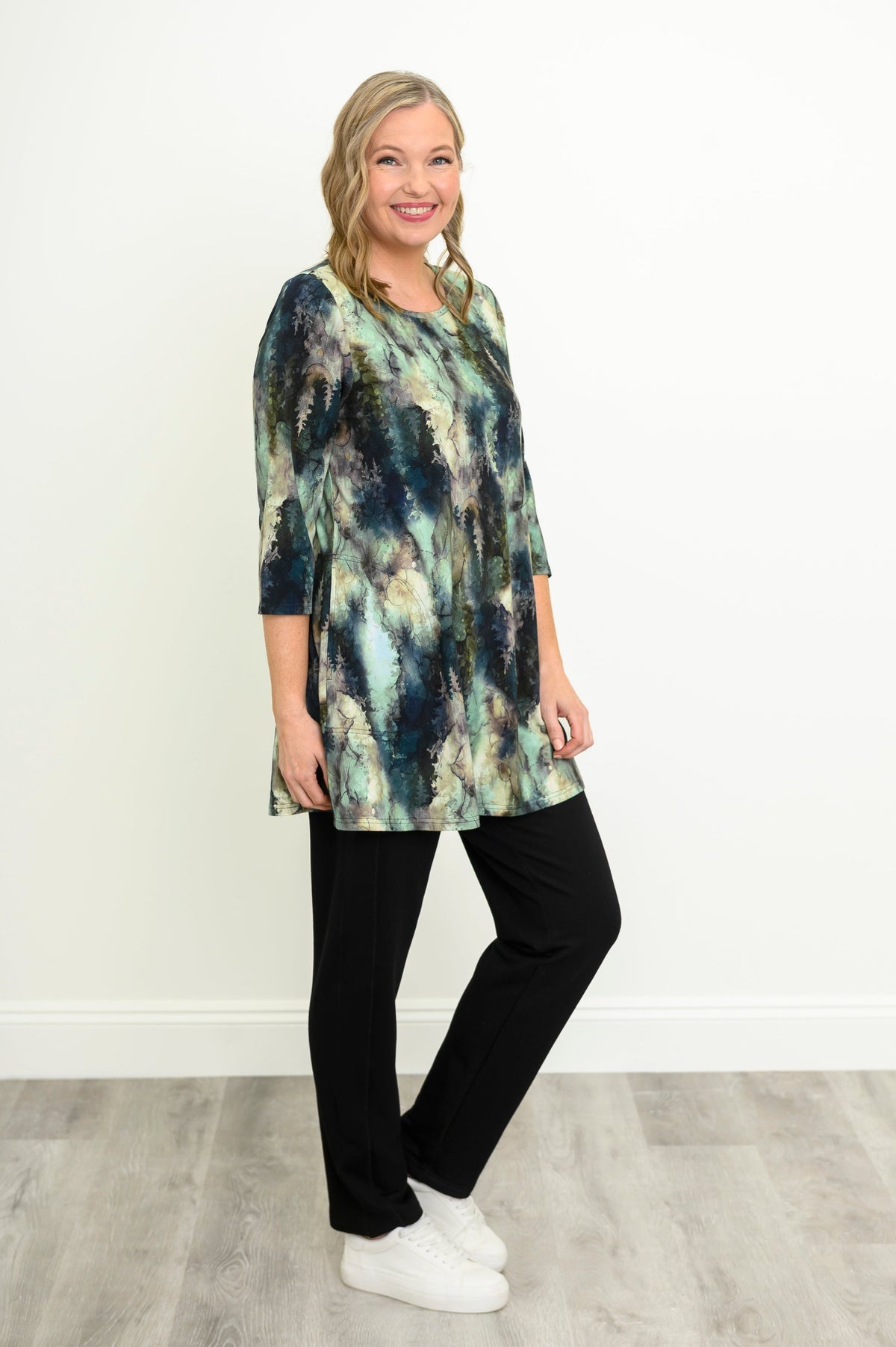 Side-facing view of a female model wearing a blue-green watercolor forest print A-line tunic with 3/4 sleeves with black pants and white shoes. Against a white wall and gray floor