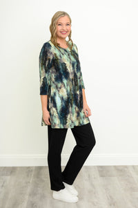 Side-facing view of a female model wearing a blue-green watercolor forest print A-line tunic with 3/4 sleeves with black pants and white shoes. Against a white wall and gray floor
