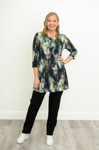 Front view of a female model wearing a blue-green watercolor forest print A-line tunic with 3/4 sleeves with black pants and white shoes, hand on hip. Against a white wall and gray floor