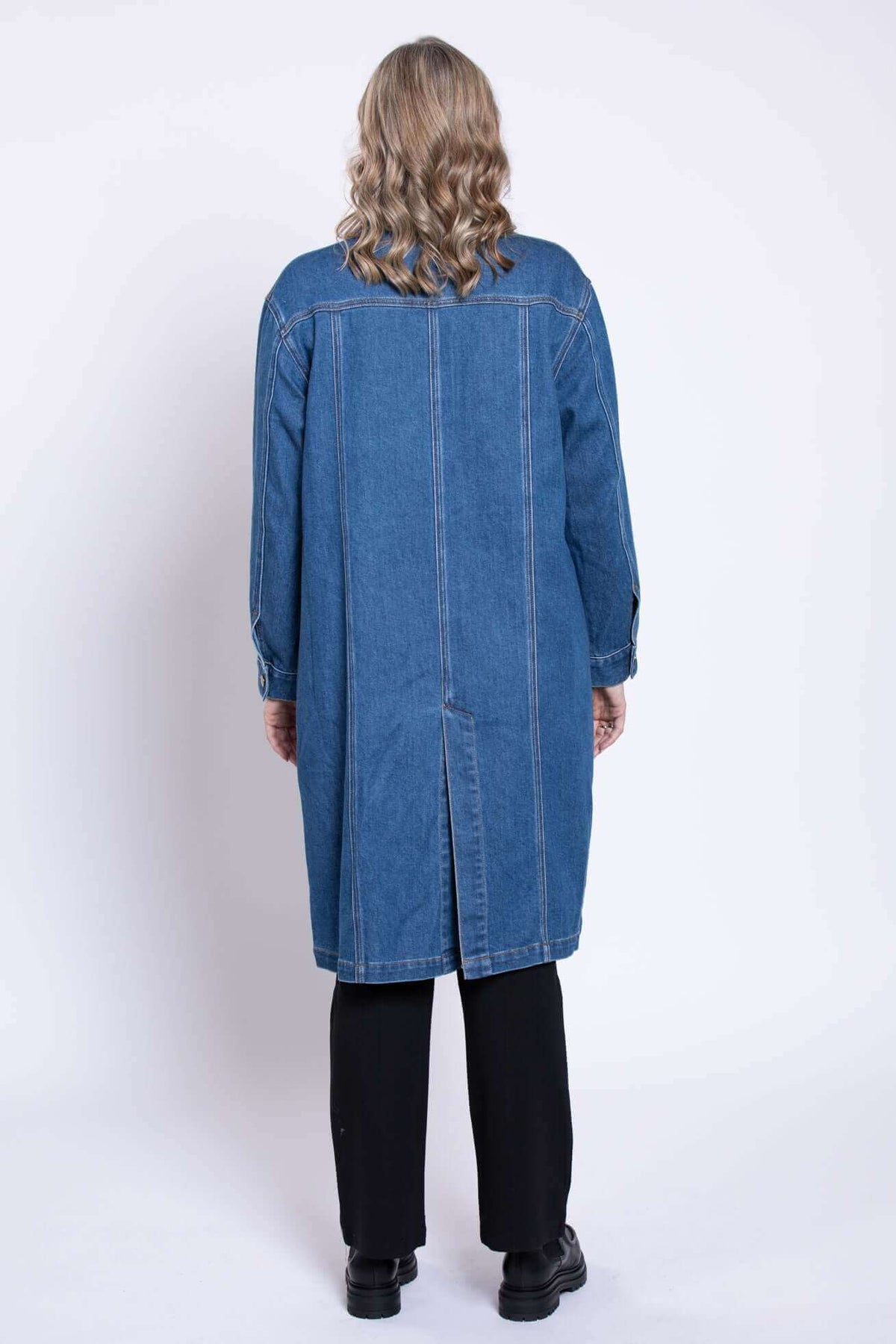 Back view of a woman wearing a knee length dark blue denim duster jacket in studio with black shoes.