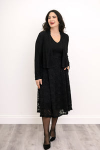 Female model smiling and posing in lace overlay dress styled with a black shrug and black heels, against white wall and gray floor