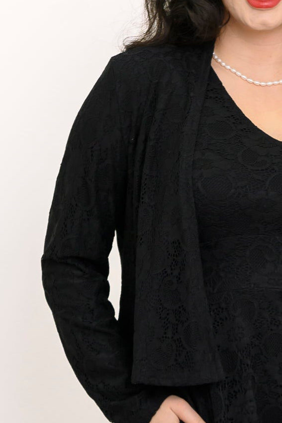 A close-up of a female model wearing a black lace cardigan over a matching black lace top, accessorized with a pearl necklace against a plain white background.