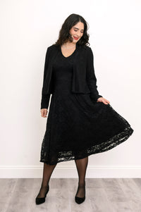 Female model playing with the hem of her black lace flared dress, smiling, against white wall and gray floor