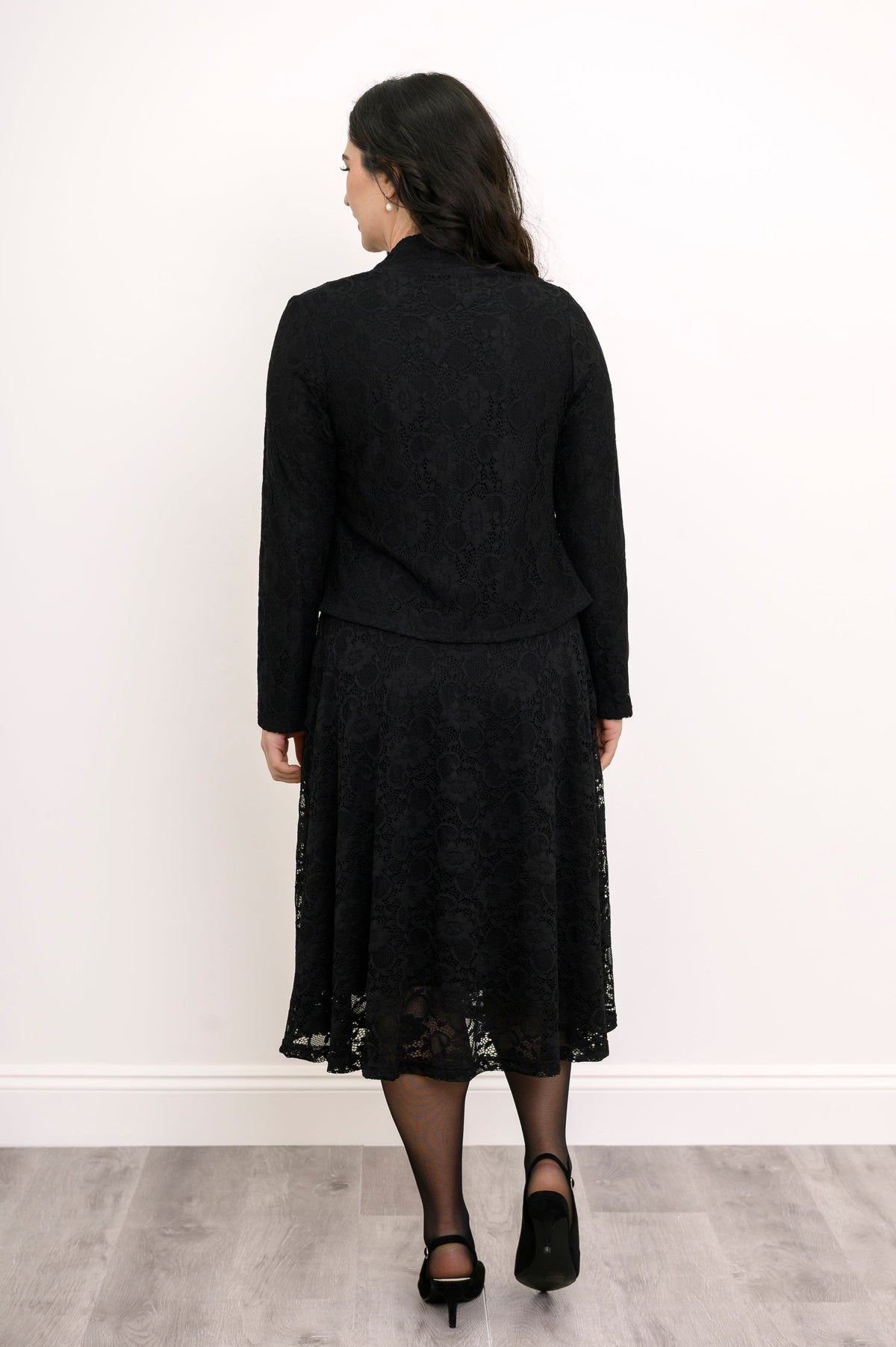 Back view of female model standing in formal black lace outfit featuring a matching cardigan and dress, against white wall and gray floor