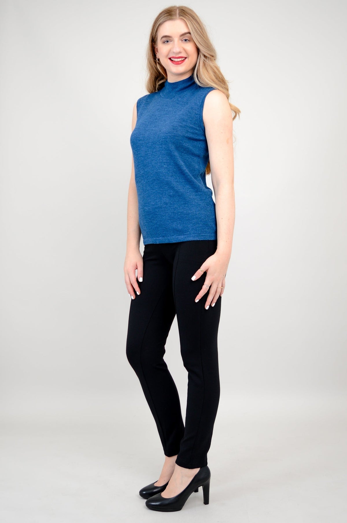 Side profile view of a woman wearing a sleeveless blue mock neck sweater showing slim fit construction.