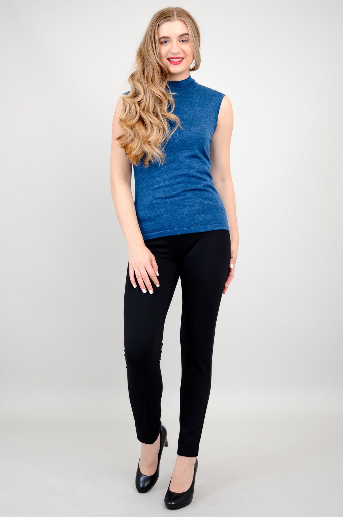 Full body image of a smiling woman modeling a bright blue sleeveless mock neck top and black trousers.