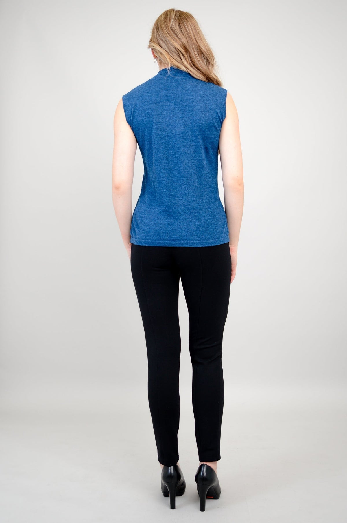 Back view of a woman wearing a sleeveless blue mock neck merino wool sweater with black trousers.