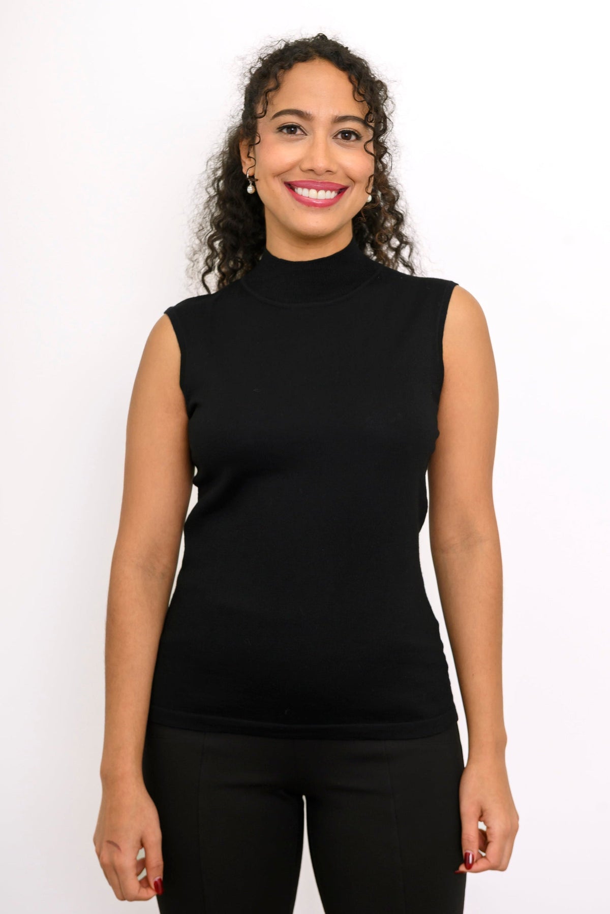 A woman with curly dark hair wearing a black sleeveless mock neck top and black pants, standing against a plain white studio background.