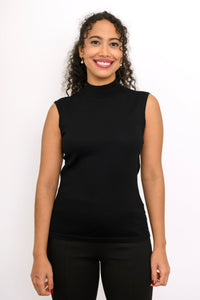 A woman with curly dark hair wearing a black sleeveless mock neck top and black pants, standing against a plain white studio background.