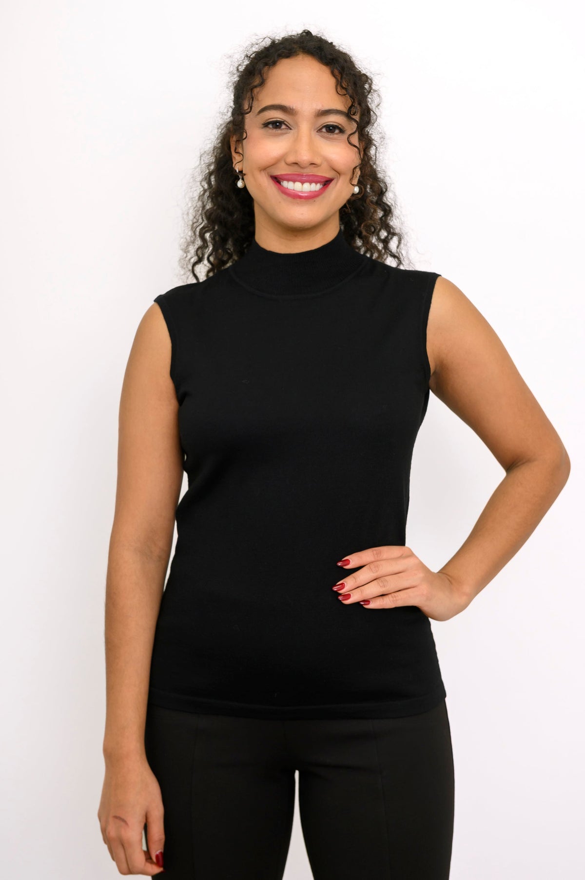 Full length seated shot of a woman in a black sleeveless mock neck sweater and matching black pants.