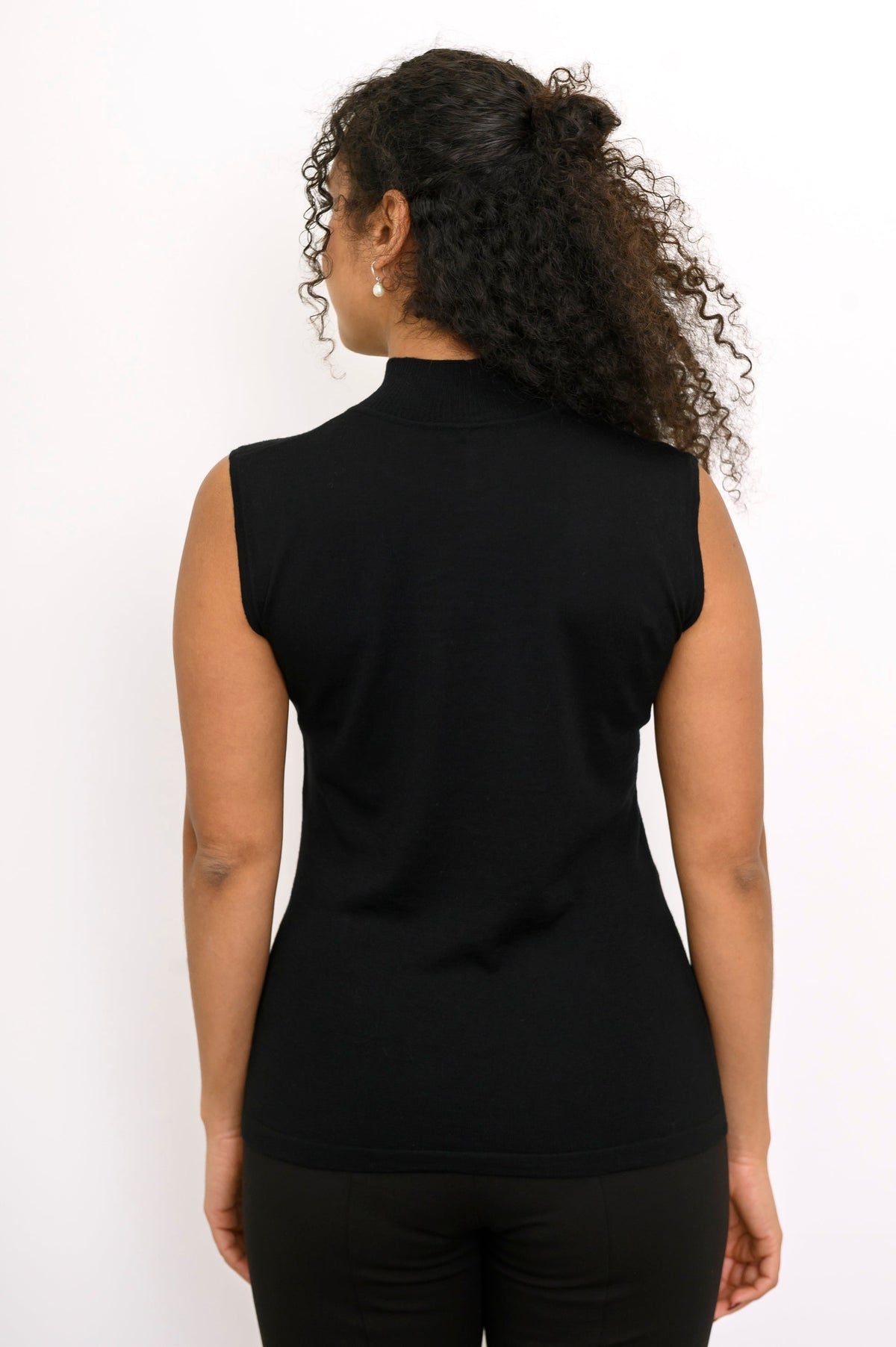Back view of a woman wearing a black sleeveless merino wool mock neck top with black trousers.