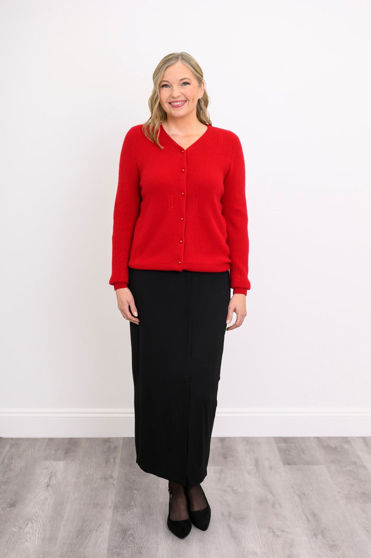 Full length view of a woman wearing a red v-neck wool cashmere cardigan paired with a midi black skirt.