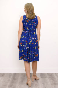 Back view of woman wearing a blue floral dress and sandals against a white wall 