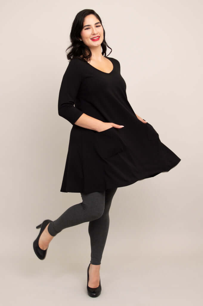 Side view of model twirling in black A-line tunic dress and grey leggings