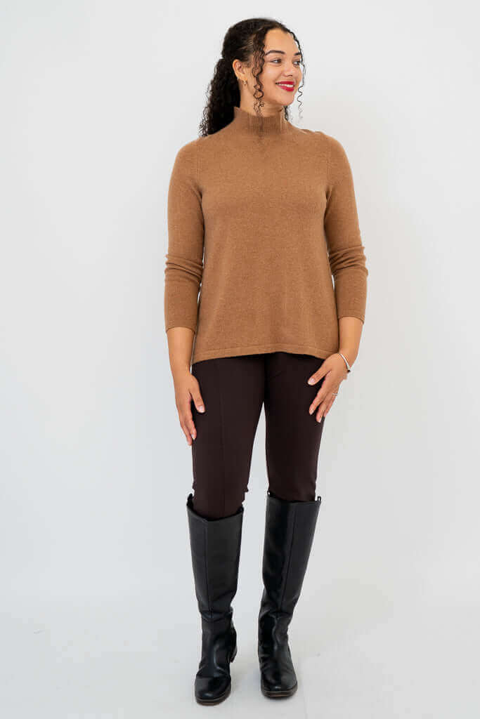 Woman in camel brown mock neck sweater with dark pants and black boots, facing slightly right