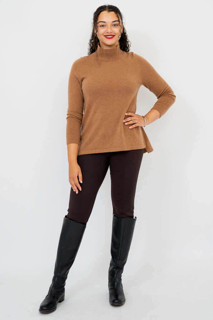 Full body view of a woman in a camel brown mock neck sweater, dark pants, and black boots
