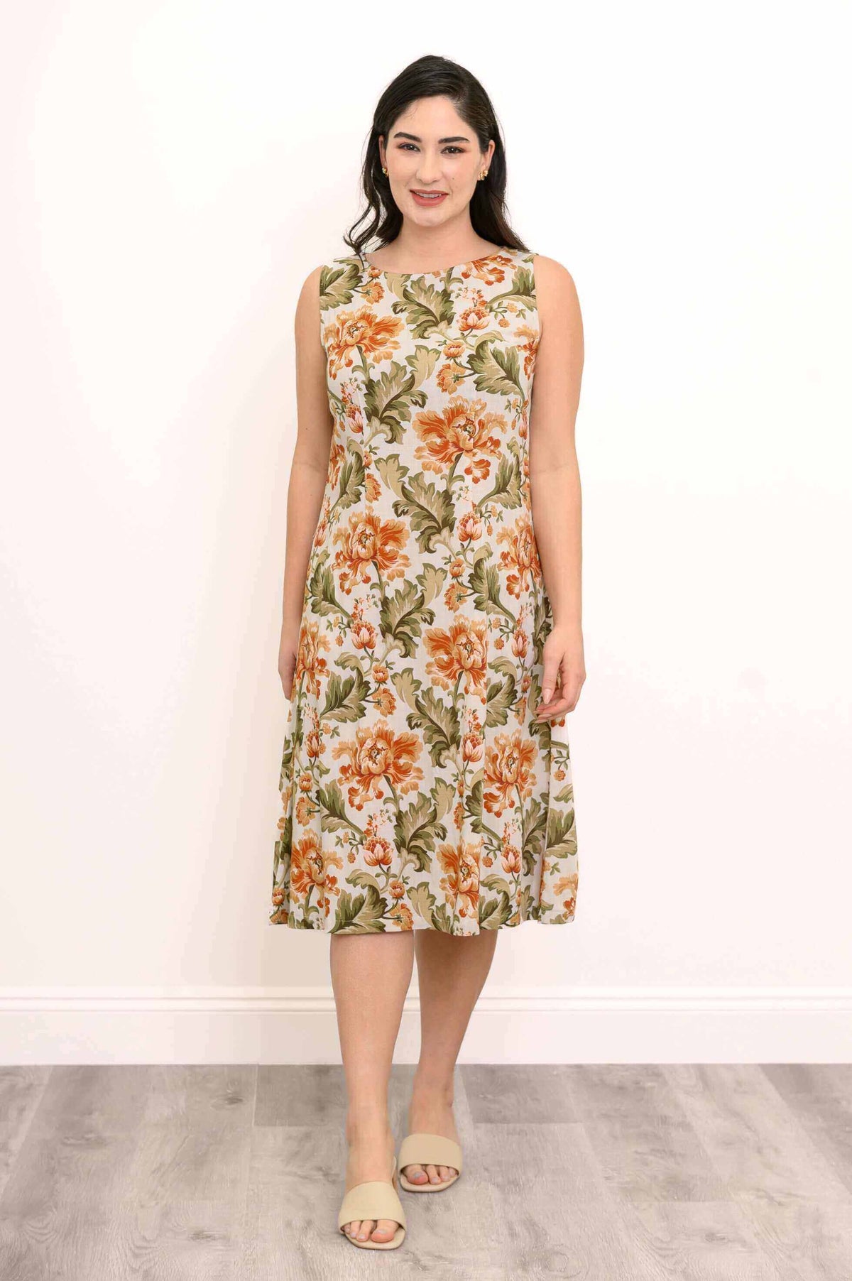 Woman wearing sleeveless floral dress in orange and green print, standing against a white wall