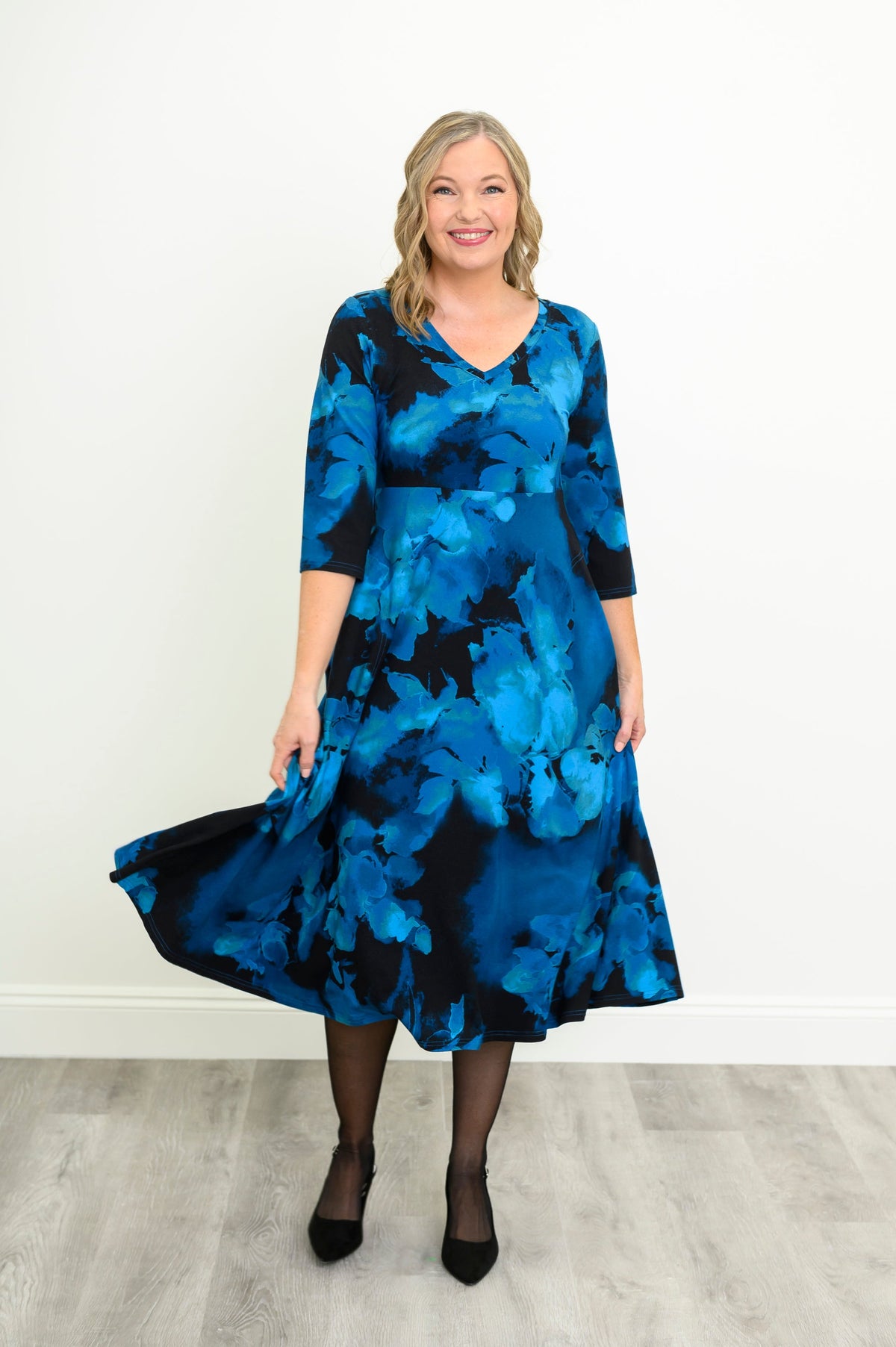 Front view of a female model wearing a blue patterned bamboo cotton midi dress styled with simple black heels, against a white wall and gray floor