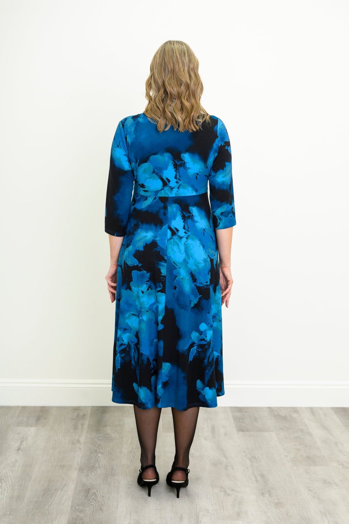 Back view of a female model wearing indigo patterned A-line midi dress highlighting the fluid skirt and full coverage, against a white wall and gray floor