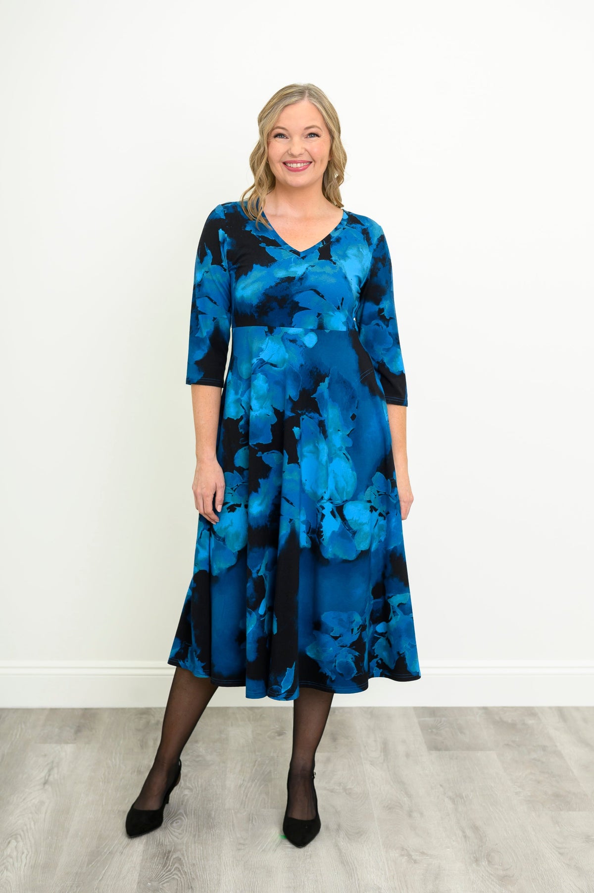 female model wearing softly draped blue and black patterned V-neck midi dress, shown from a slight side-angle, against white wall and grey floor