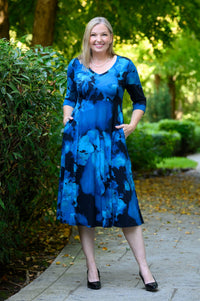 female model stands on a paved path in a park wearing a blue and black floral midi dress with three-quarter sleeves and side pockets. The background consists of lush green trees and bushes.